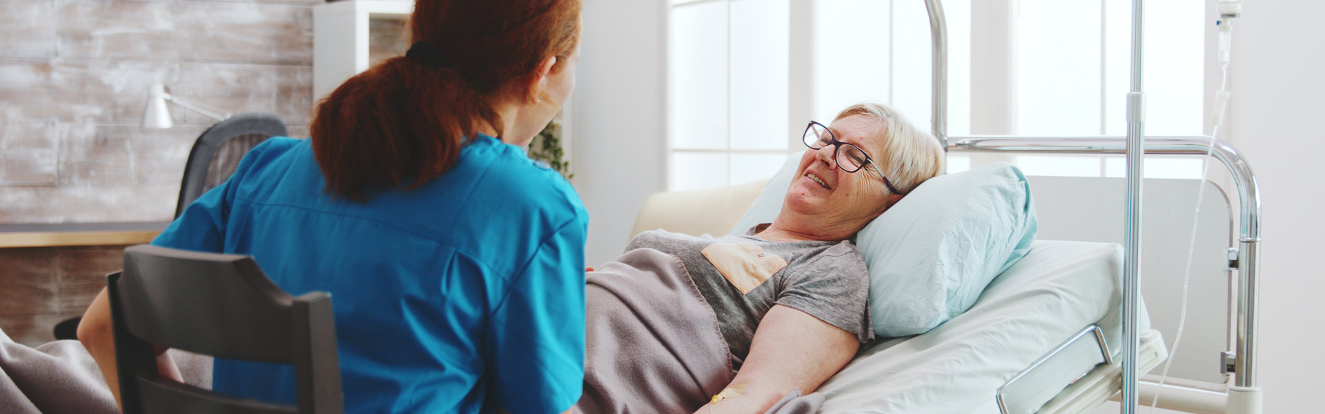 In bright retirement home old lady lying in hospital bed talks with a female caregiver